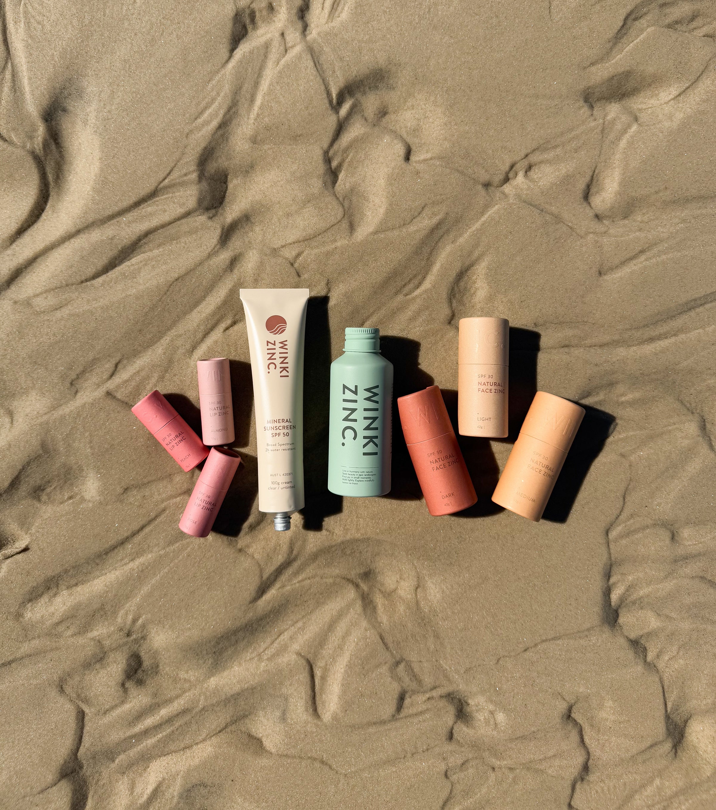 Winki Zinc natural mineral sunscreen, tinted face and lip zinc sticks and cleanser laying on sand.