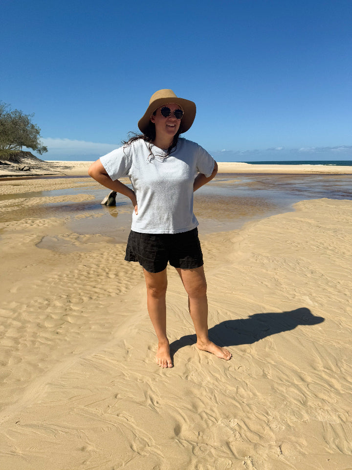 Woman wearing Arius Australian made The Vera T-Shirt in dusty blue Good Earth Cotton while at the beach standing smiling in the sand. 