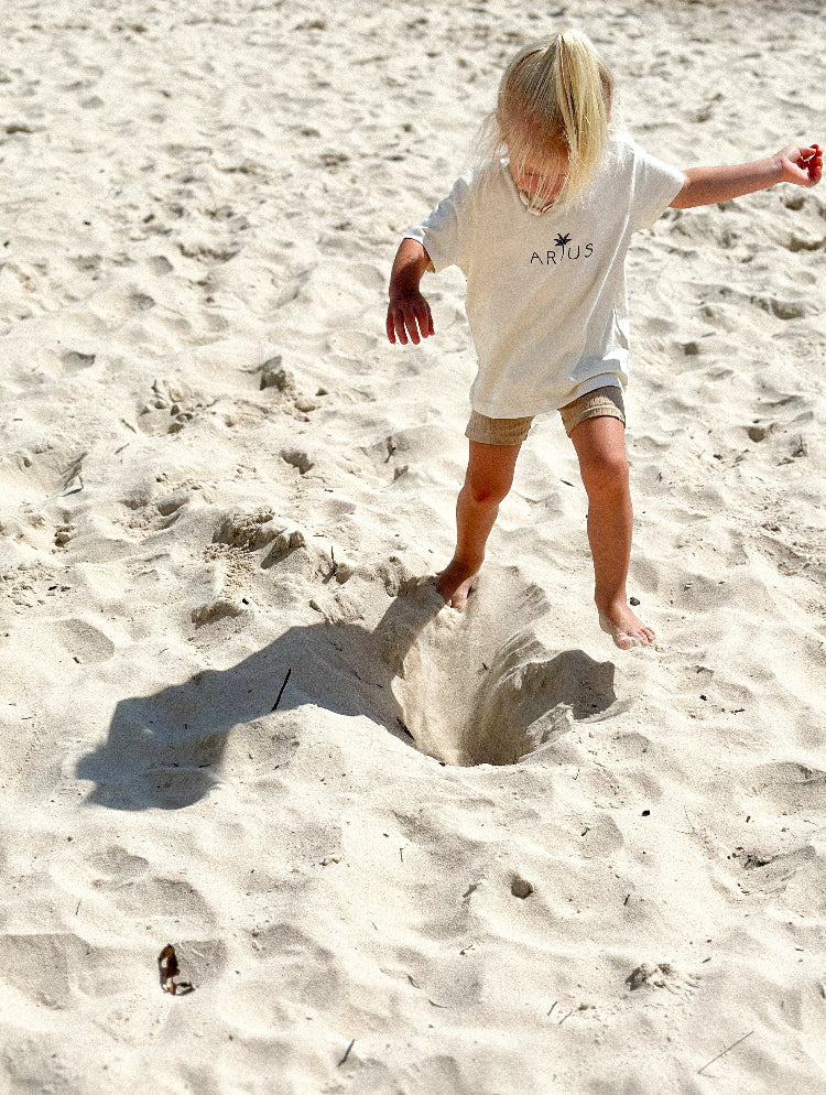Child wearing Baby Arius Organic Cotton T-Shirt in natural with palm tree logo printed in centre and child running across sand. 