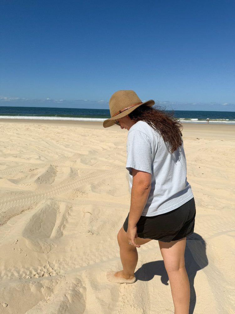 Mid-sized woman wearing The Vera Good Earth Cotton Arius T-Shirt in inclusive sizing while at the beach walking on sand. 