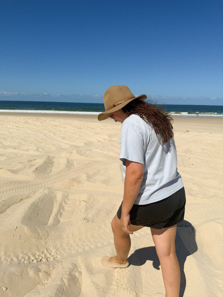 Mid-sized woman wearing The Vera Good Earth Cotton Arius T-Shirt in inclusive sizing while at the beach walking on sand. 