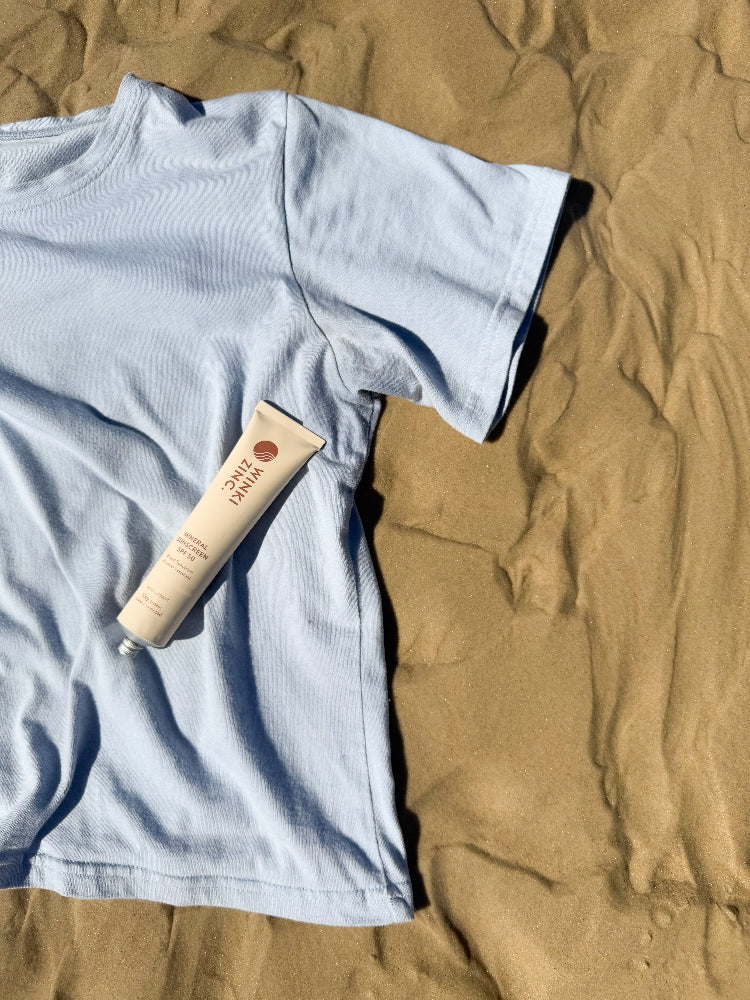 Arius The Vera T-Shirt made in Australia from Good Earth Cotton in a dusty blue colour laying on the sand at the beach with natural mineral zinc sunscreen tube laying on t-shirt. 