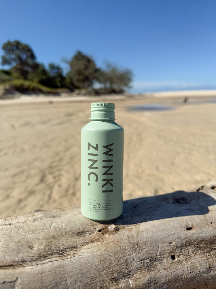 Bottle of Winki Zinc cucumber and avocado oil natural face cleanser displayed on a log of driftwood at the beach. 