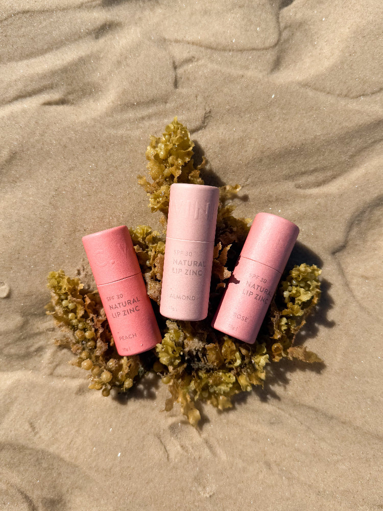 Winki Zinc natural lip zinc in three tints laying on a piece of seaweed atop sand at the beach. 