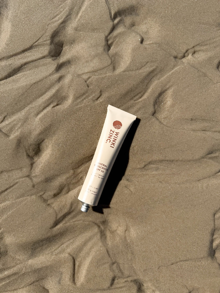 Winki Zinc Mineral Sunscreen cream coloured aluminium tube laying on the sand at the beach. 