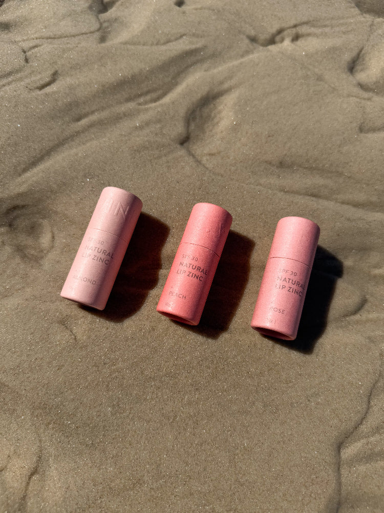 Three Winki Zinc natural lip zinc SPF 30 plastic free compostable tubes laying in the sand at the beach. 