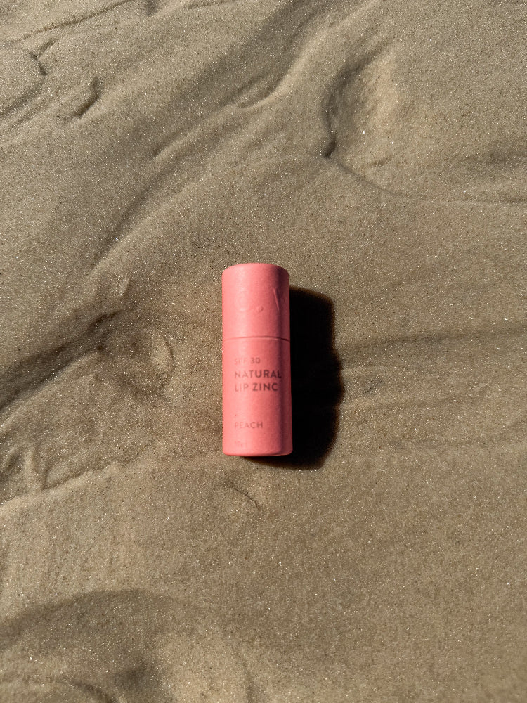 Winki Zinc natural lip zinc in peach tint plastic free tube laying on beach. 