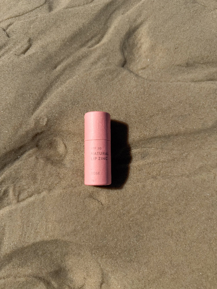 Winki Zinc SPF 30 compostable cardboard tube natural lip zinc in rose shade laying on sand. 