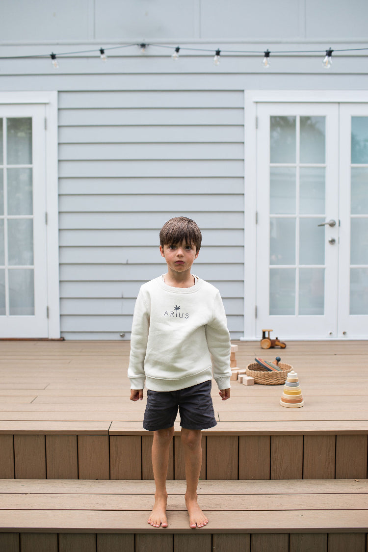Kid wearing ethically made organic cotton jumper in natural with palm tree logo while standing on deck. 