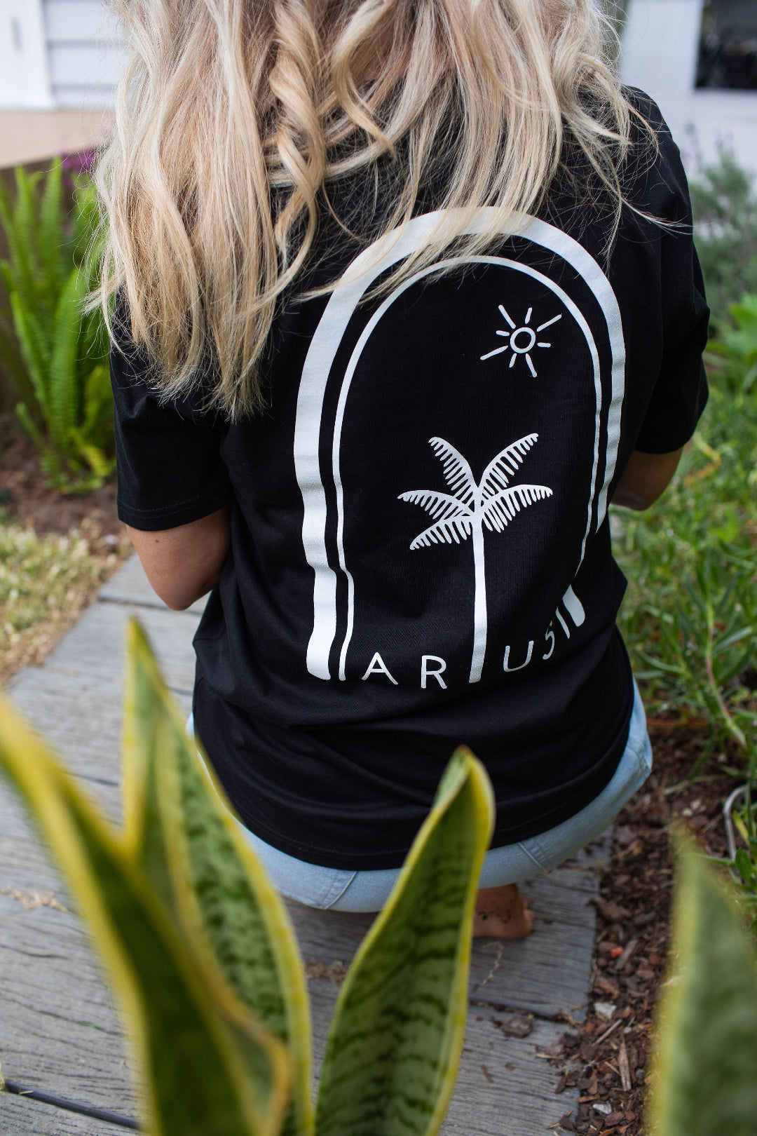 Woman wearing unisex organic cotton t-shirt in black showing back of t-shirt with large palm tree and sun print in white. 
