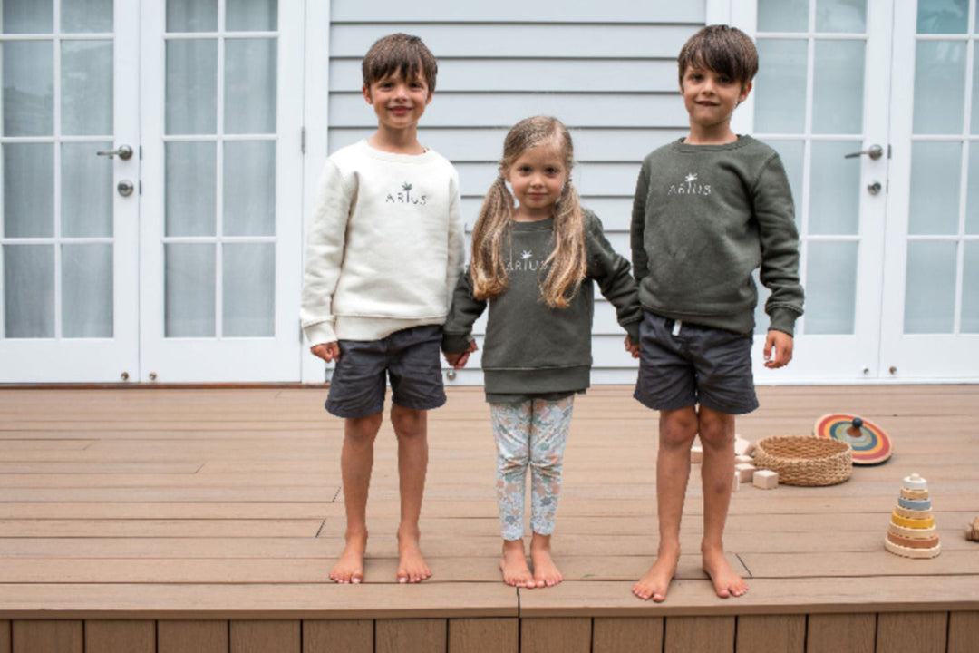 Three siblings holding hands on a deck wearing Arius organic cotton kids jumper in natural and khaki green. 