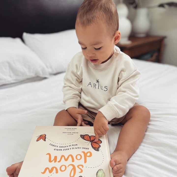 Baby sitting on bed wearing cozy organic cotton jumper in natural colour with palm tree logo reading a book. 
