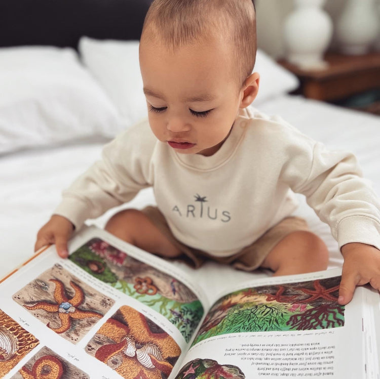 Baby reading a book on bed wearing Arius Baby Snuggler cozy organic cotton jumper in natural colour with palm tree logo. 
