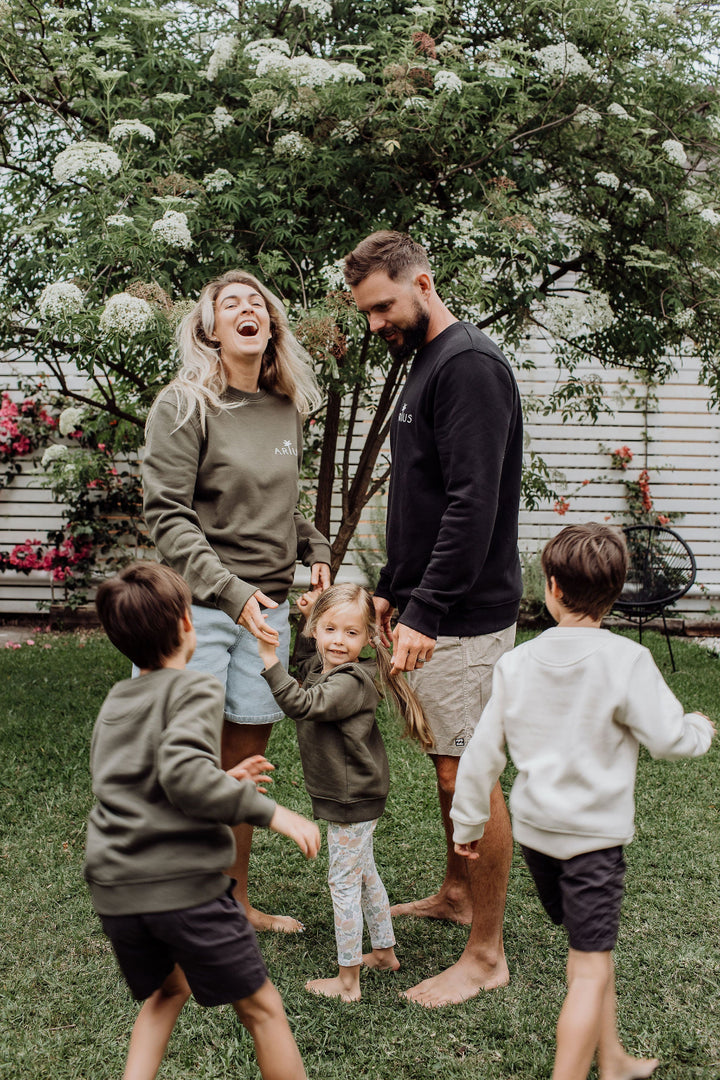 Family playing in backyard all wearing matching organic cotton Arius jumpers in natural, black and khaki green. 