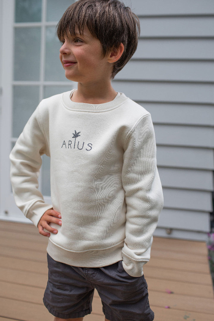 Kid wearing ethically made organic cotton jumper in natural with palm tree logo. 