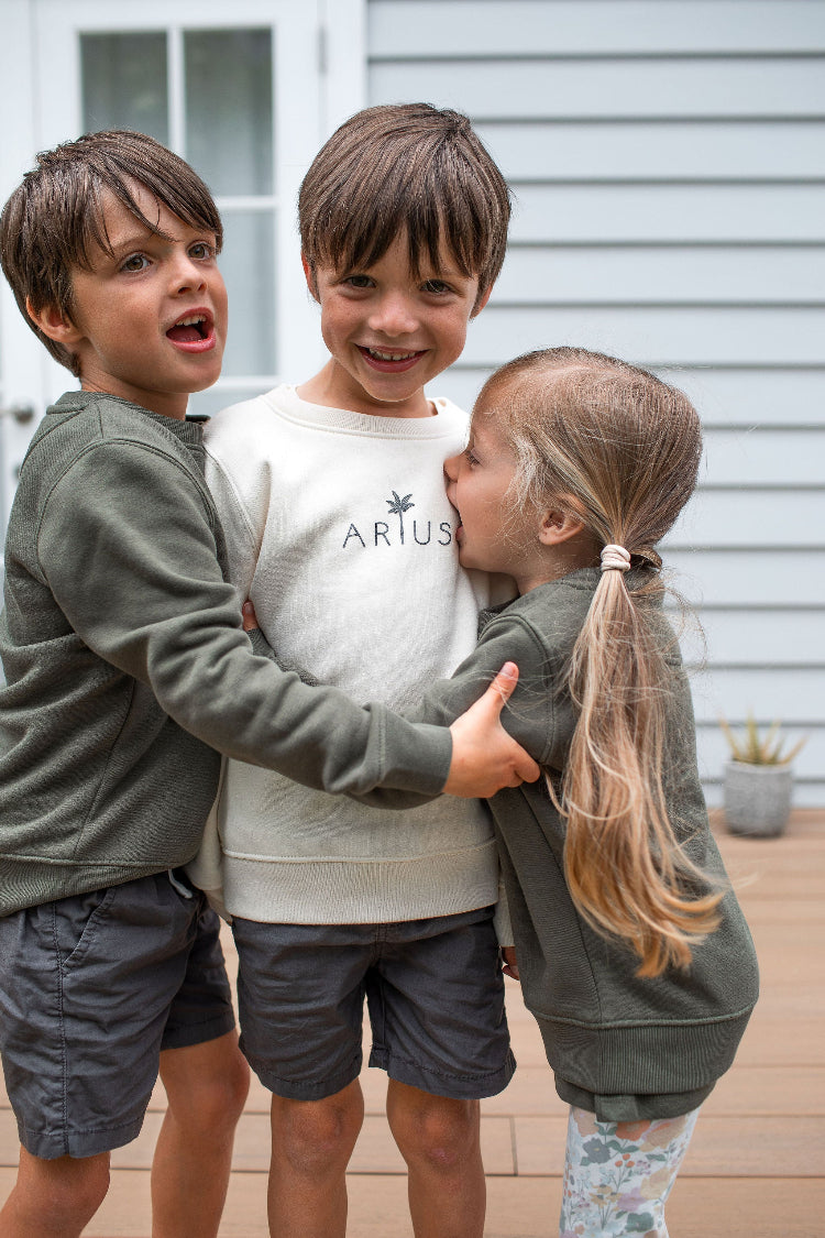 Three kids hugging all wearing Arius organic cotton kids jumpers in natural and khaki green. 
