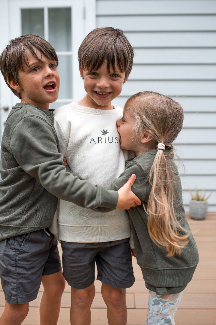 Three kids hugging all wearing Arius organic cotton kids jumpers in natural and khaki green. 
